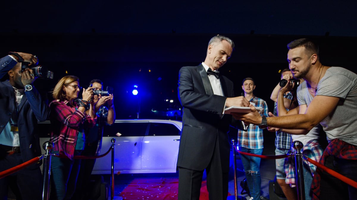 Handsome adult man in tuxedo walking on red carpet and giving autographs to fan people