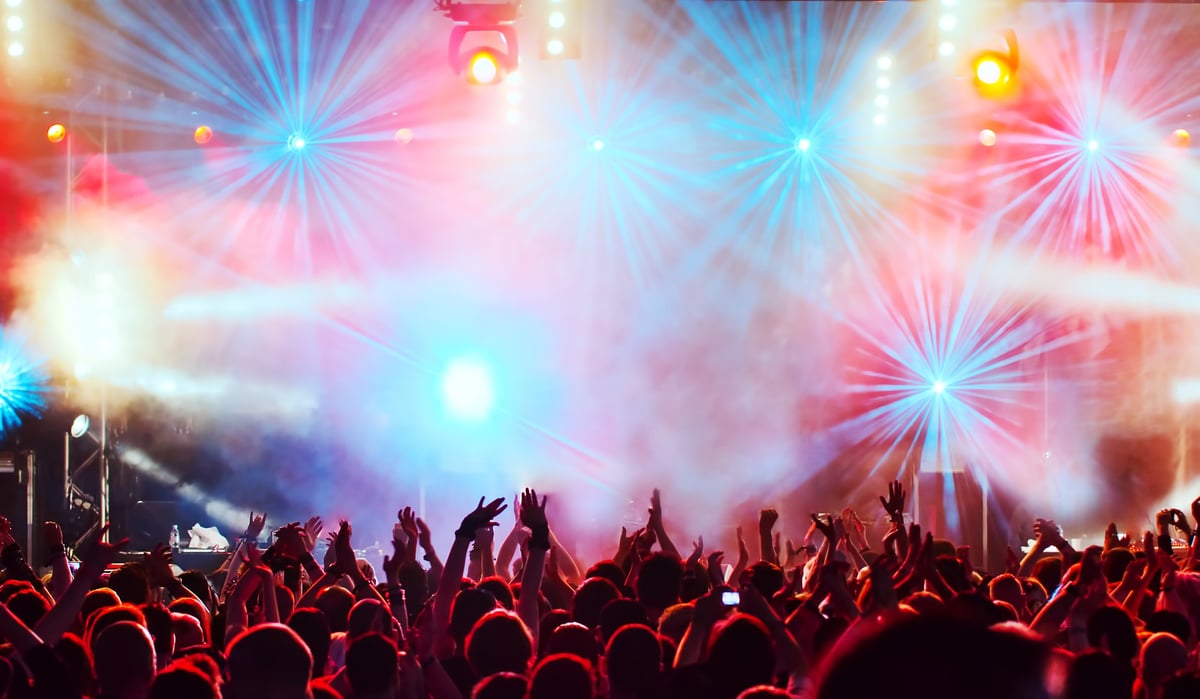 A crowd with raised hands at a concert, illuminated by vibrant red and blue stage lights and fog effects