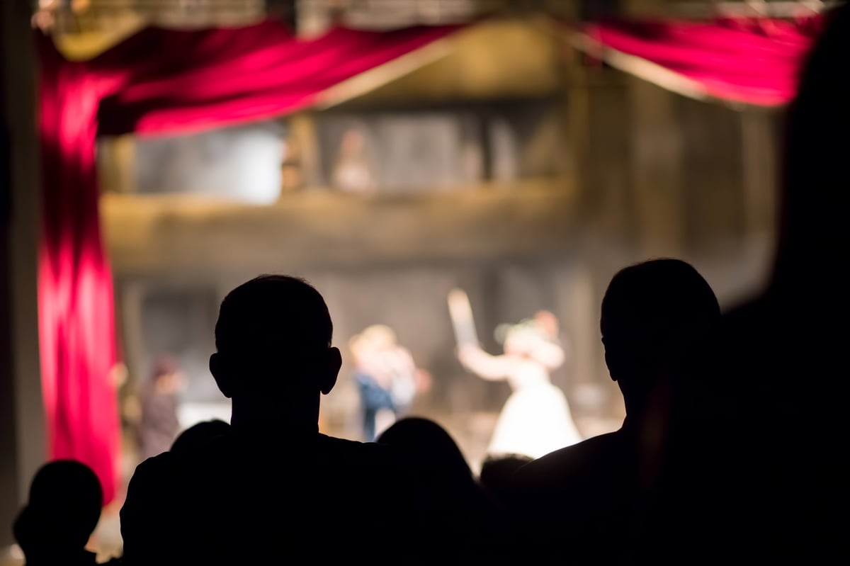 Silhouettes of audience members watching a ballet performance on stage with red curtains and dim lighting