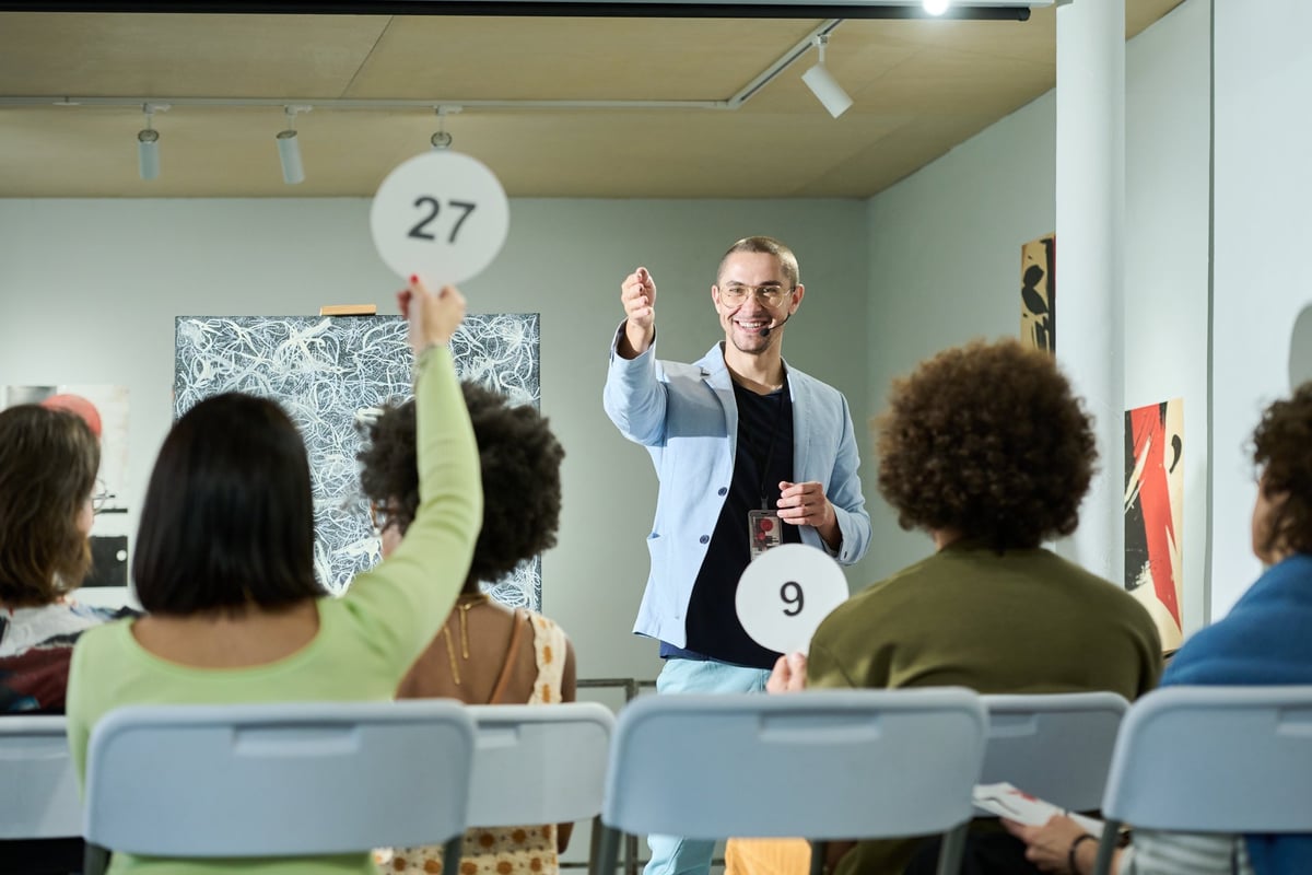 Happy young man standing in front of public raising paddles and pointing at one of female auctioneers during bidding