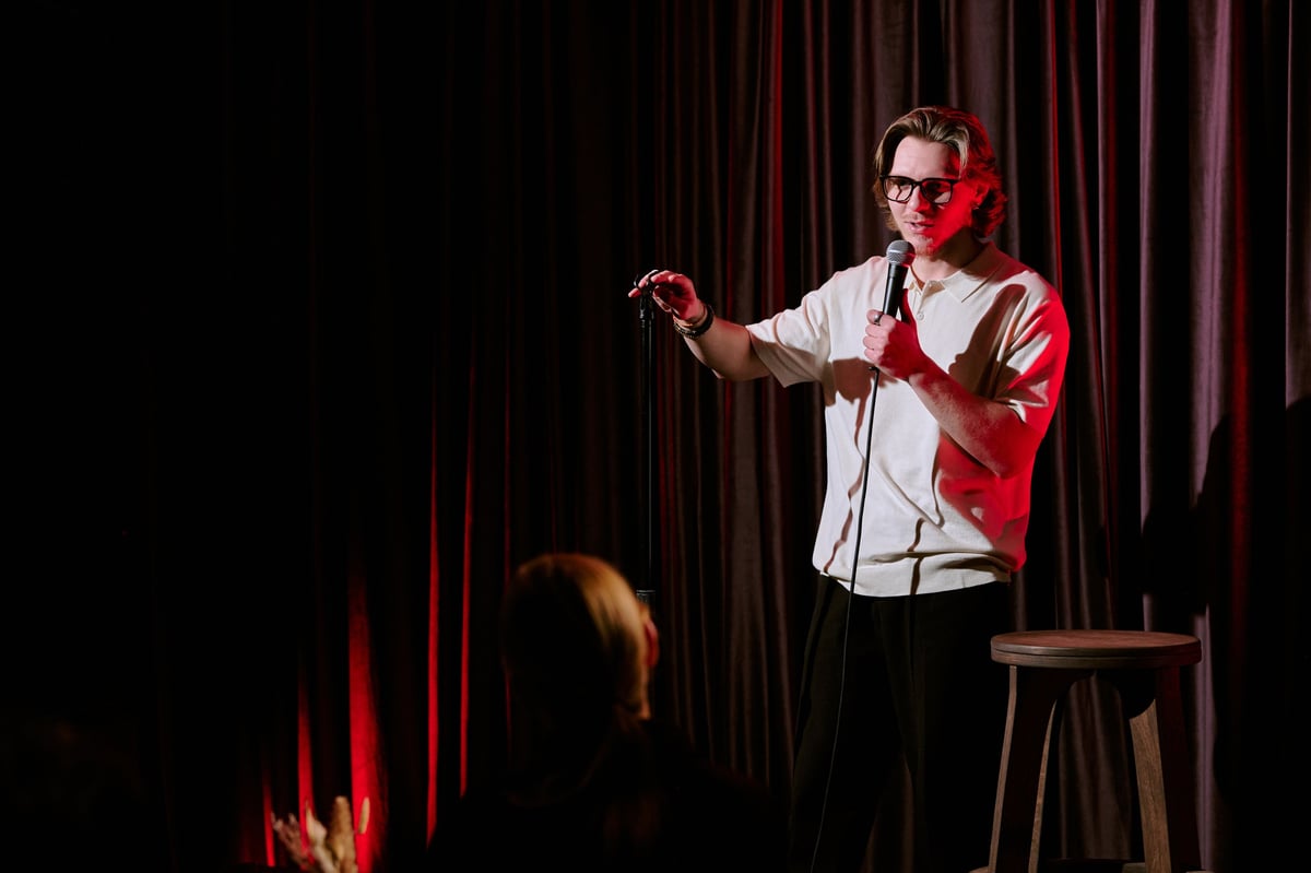 Comedian performing stand-up routine on stage, holding microphone and engaging with audience under dramatic lighting conditions