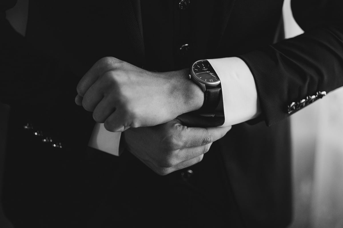 A man is wearing a black suit and is looking at his watch. The watch is black and has a silver band.