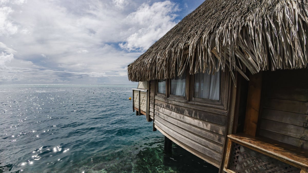 A serene overwater bungalow with a thatched roof overlooks the sparkling ocean under a partly cloudy sky, evoking tropical paradise vibes