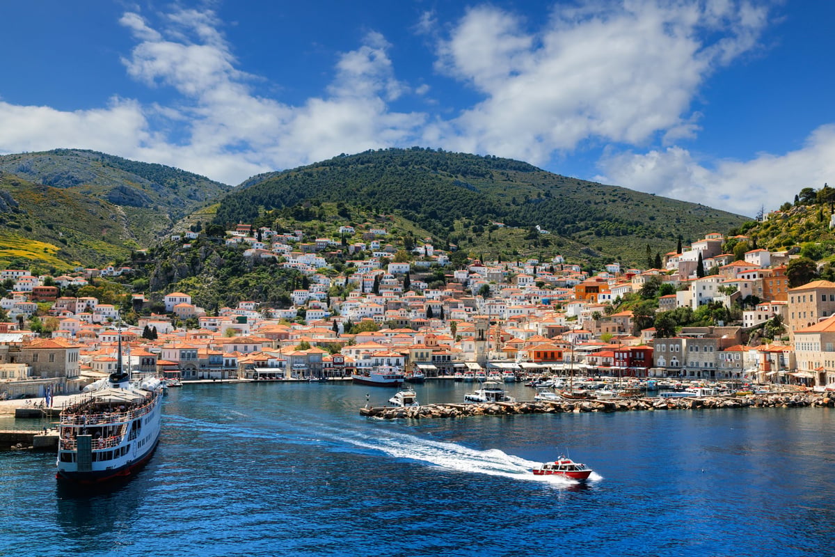 Vivid view of Hydra Island harbor, Greece, with turquoise waters, docked boats, and traditional hillside houses under a bright Mediterranean sky.