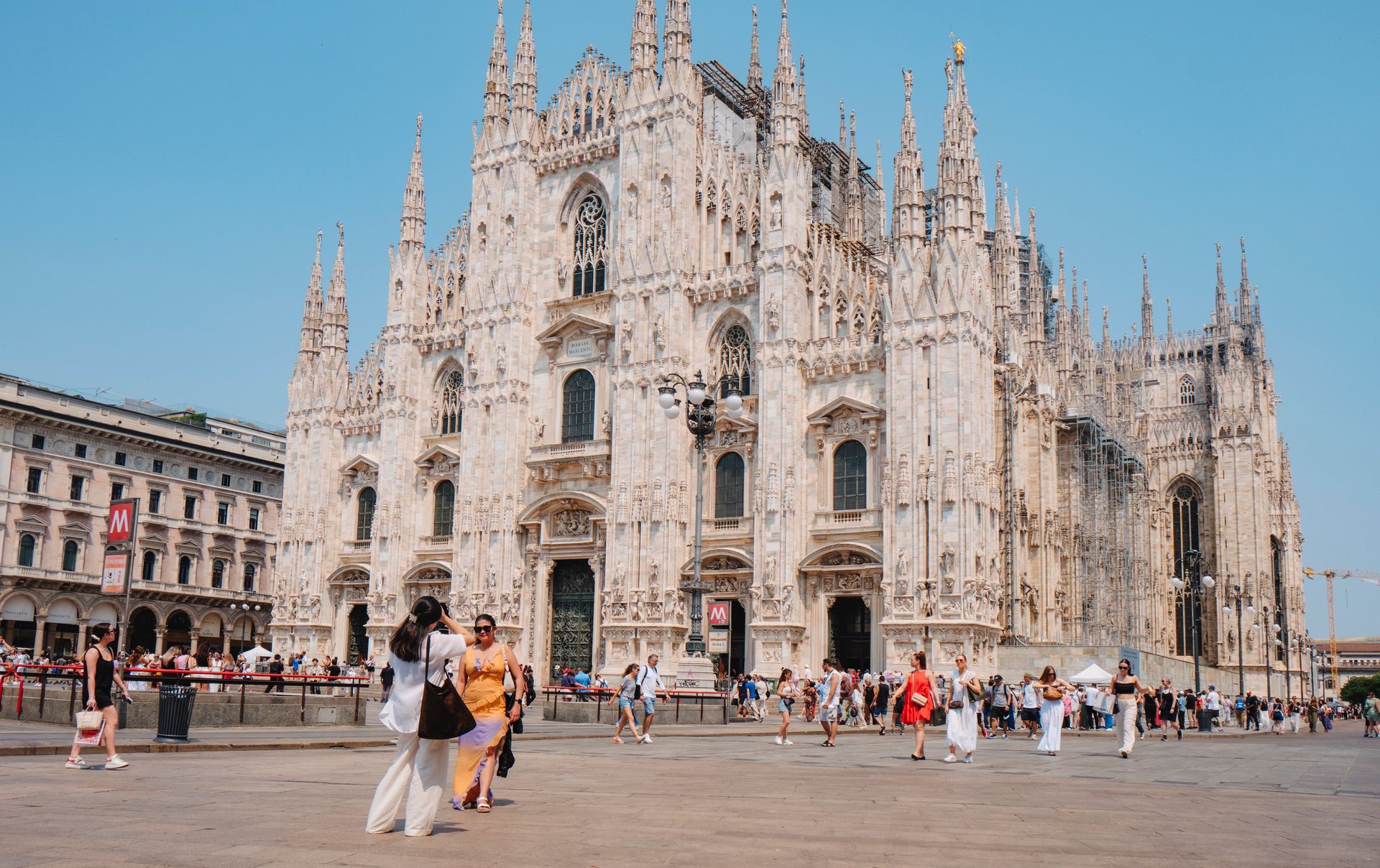 Milan Cathedral with tourists in the square under a clear sky, showing the cathedral facade and Gothic details