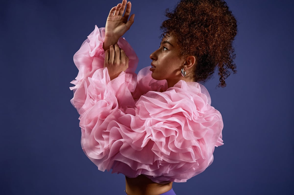 Vertical shot of young adult multiethnic woman posing in profile with arms raised, wearing voluminous ruffled top, curly hair styled up