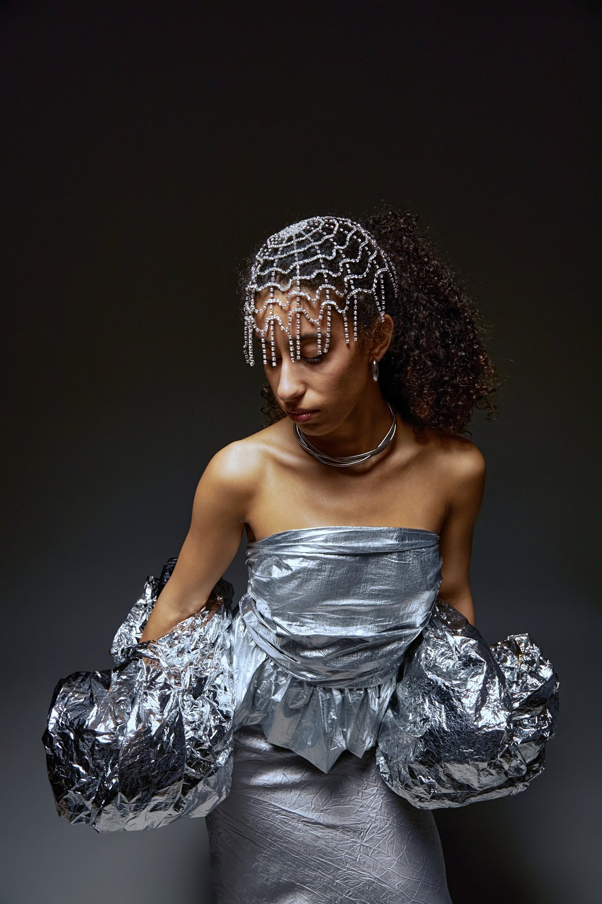 Vertical shot of young adult multiethnic woman posing in metallic strapless dress with elaborate beaded headpiece, looking down during fashion photoshoot, curly hair styled back, arms in shiny fabric