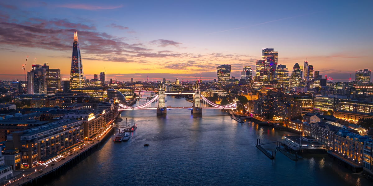 A stunning aerial panoramic view of London at twilight, featuring the Tower Bridge illuminated over the River Thames and the modern City skyline