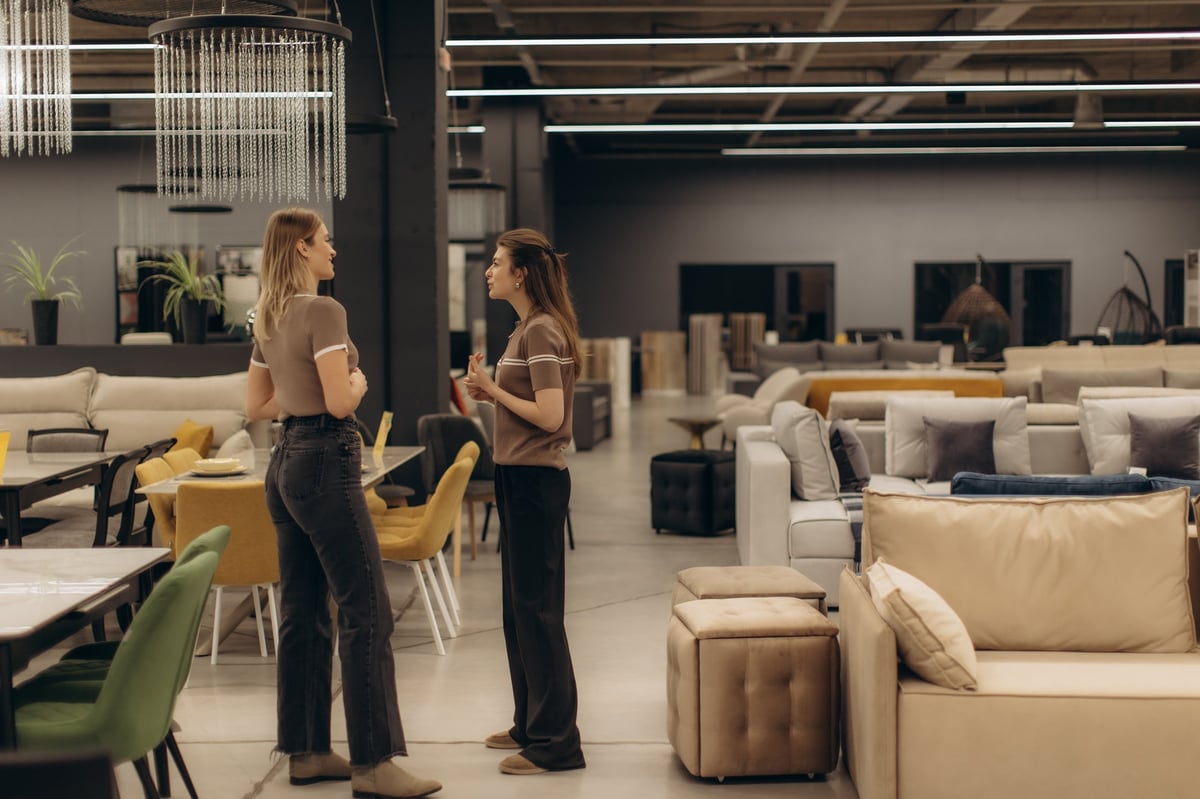 Two individuals engaged in conversation within a spacious, stylish furniture showroom, surrounded by a variety of sofas, chairs, and tables, showcasing modern interior design and decor elements