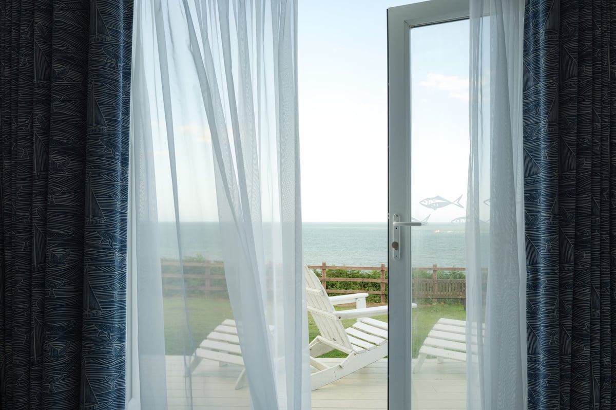 View of a cliff top seaside lodge looking out from the living room onto an open patio door. Wooden furniture is for guests to enjoy while looking at the sea.