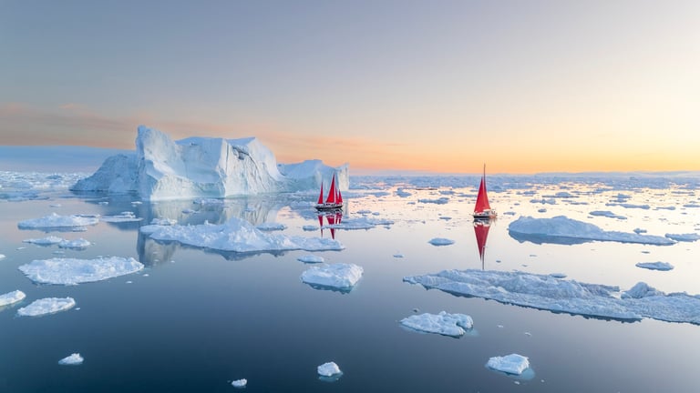 Sailboats navigating icy polar waters at sunset with towering icebergs