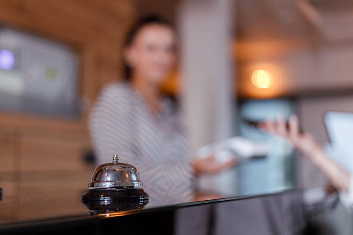 A hotel front desk features a bell while a staff member assists a guest with check-in