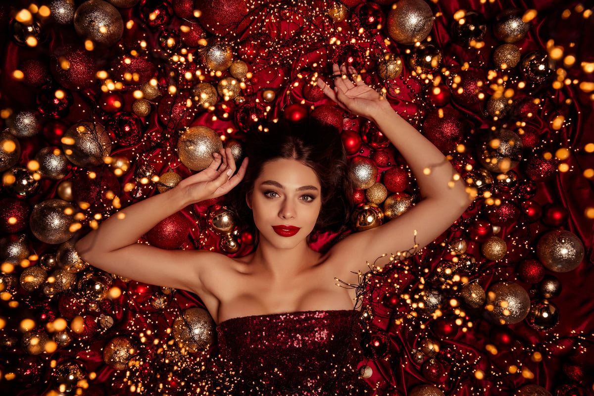 A beautiful model lies among Christmas decorations and festive lights wearing a red sequin dress