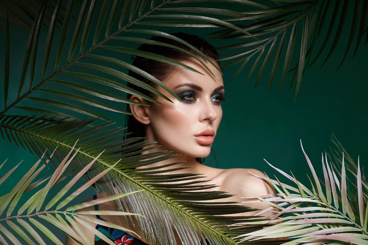 Model showcases striking makeup and fashion while posing amongst lush green palm leaves