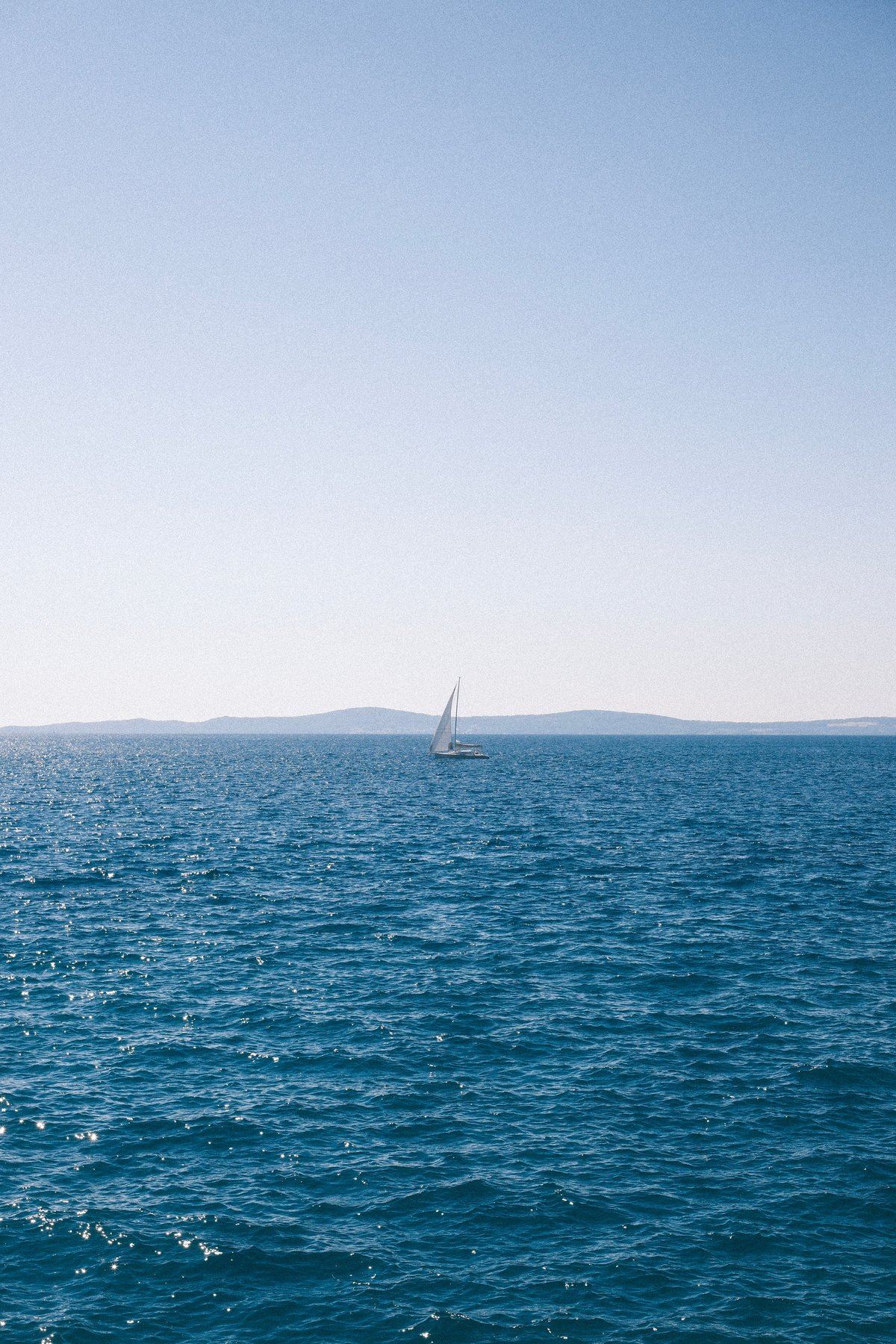 Small sailboat on a vast blue ocean with distant hills under a clear sky