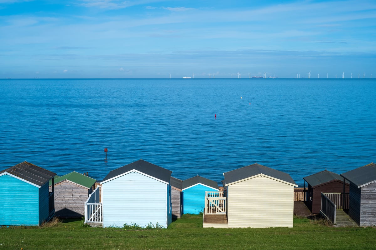 Wooden beach huts along a grass verge by a pebble beach with beautiful blue sea