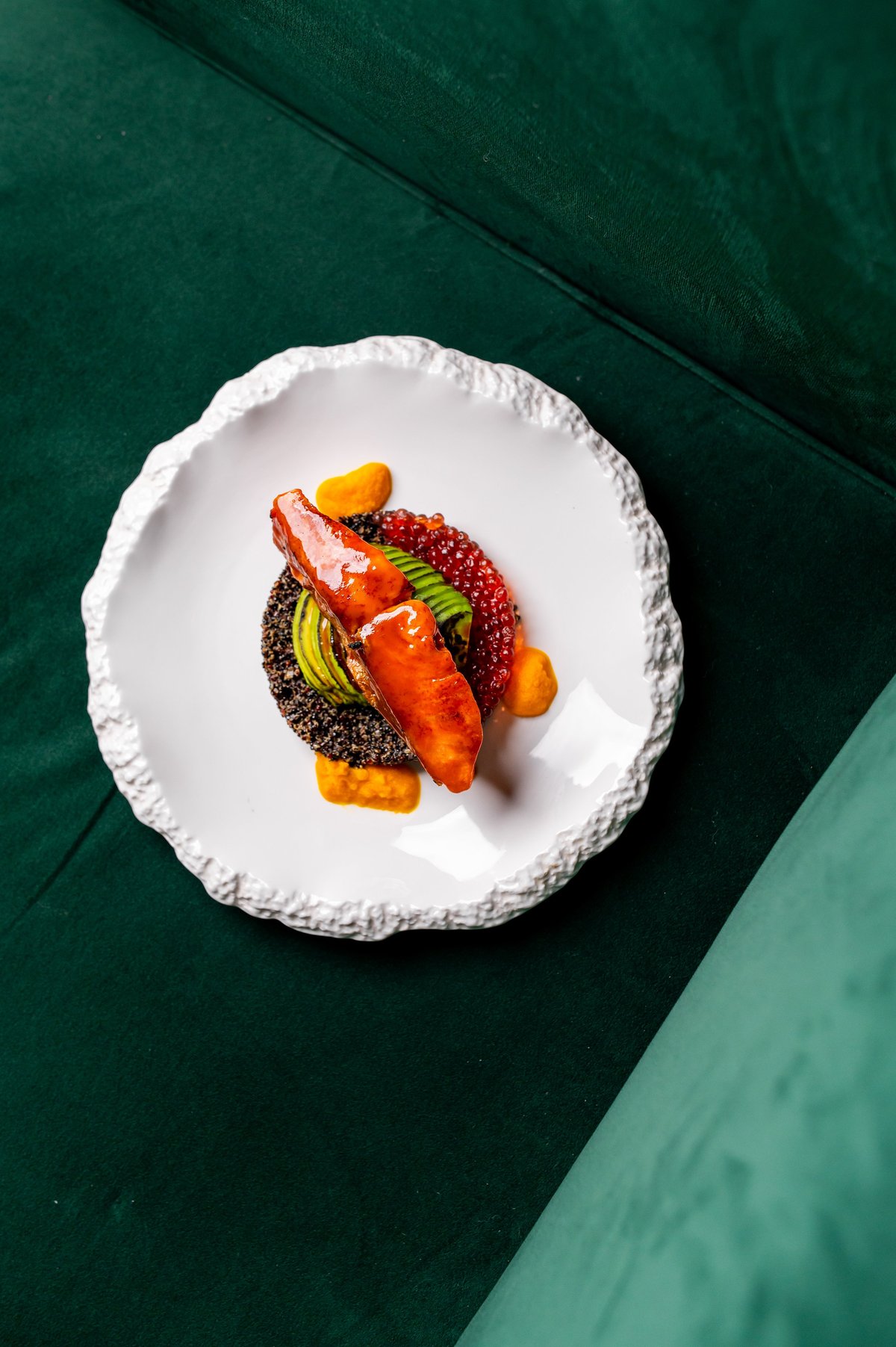 Gourmet dish with plated salmon, red caviar, avocado, and sauce on a modern white plate. Fine dining meal served on a minimalist plate against a deep green background