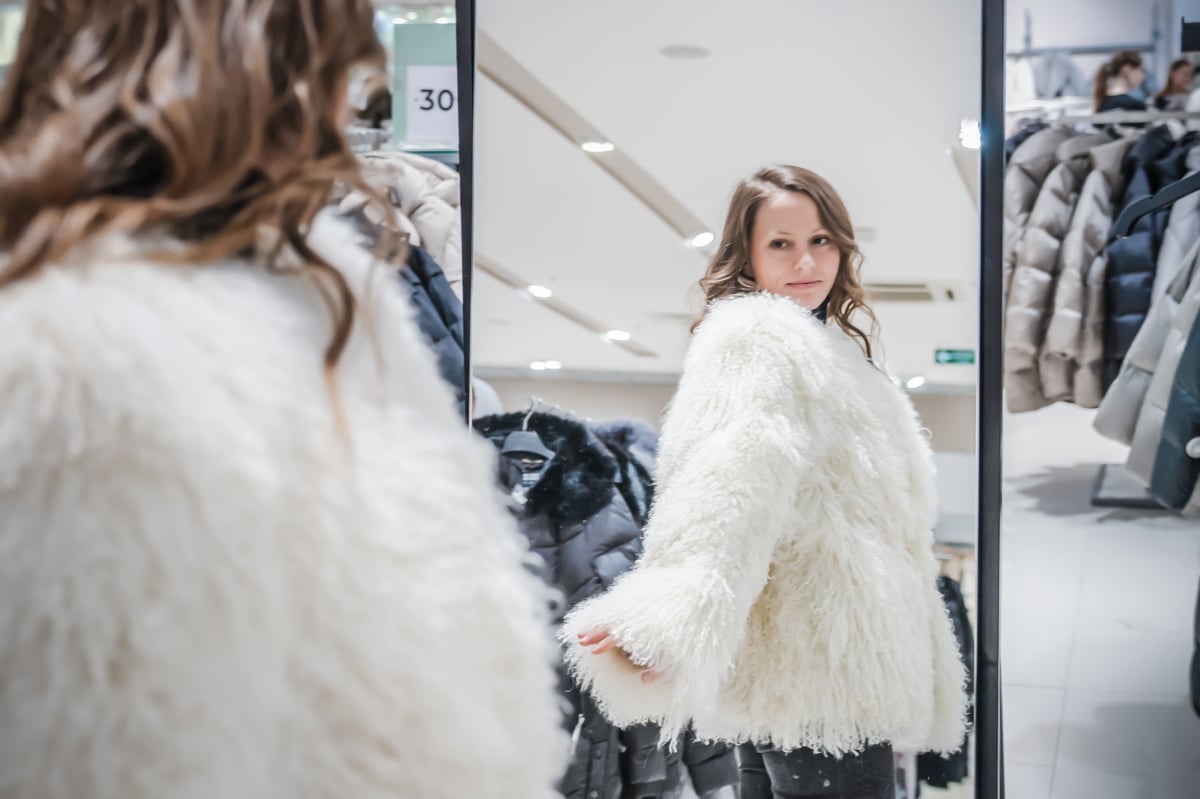 Young woman trying on stylish white sheepskin coat in clothing boutique checking reflection