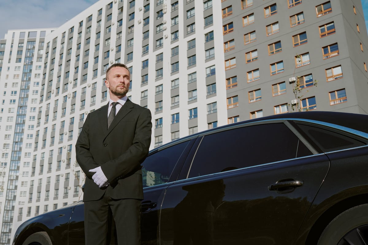 Caucasian middle aged man standing in front of black car outside modern apartment building, wearing suit and gloves, looking away with serious expression, acting as security guard
