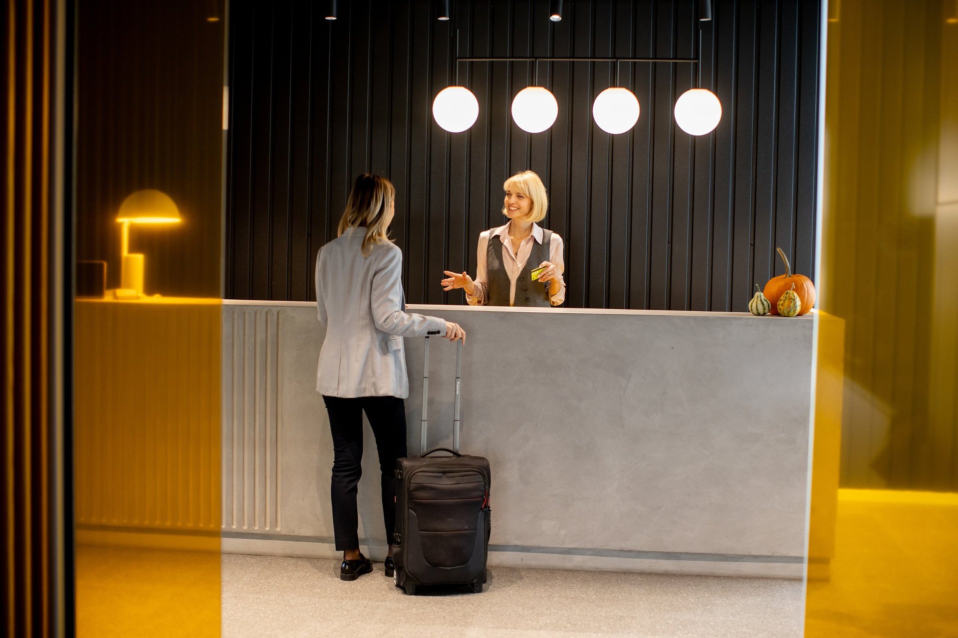 A guest arrives at the hotel reception, where a smiling staff member greets her warmly. The modern decor and warm lighting create a friendly atmosphere for visitors.