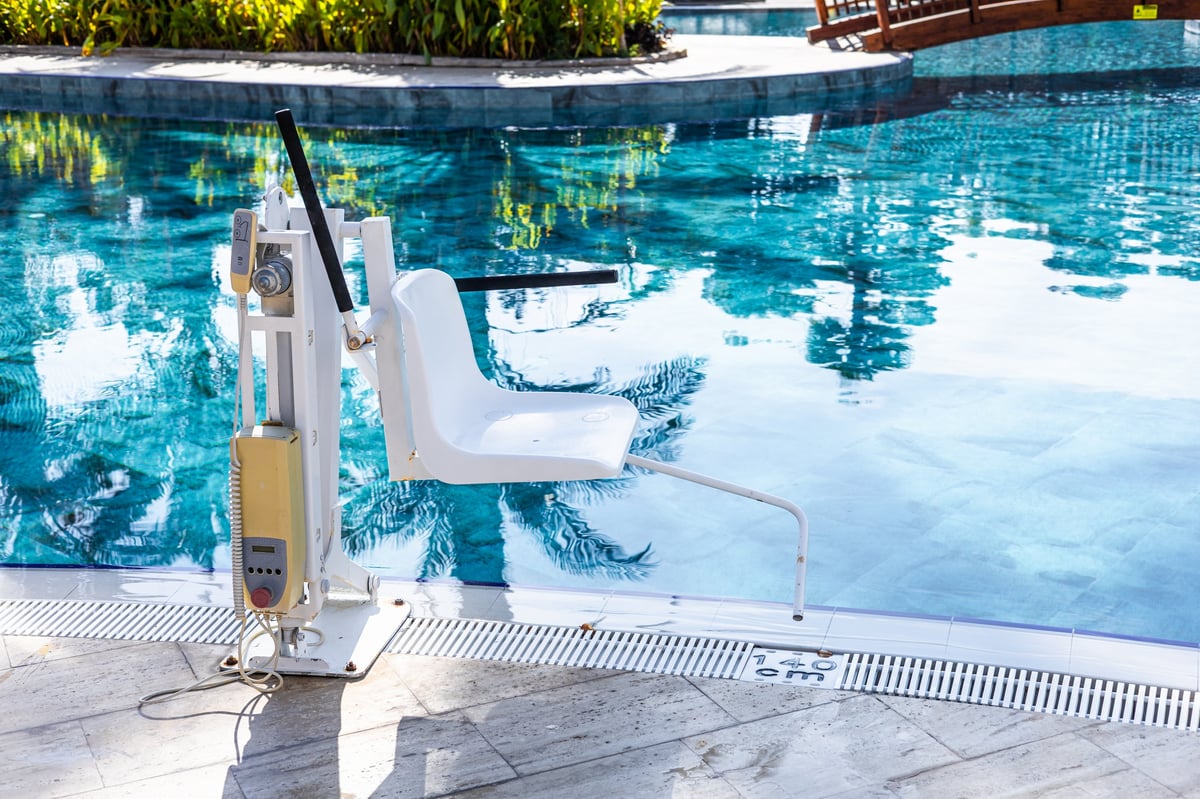 A poolside scene featuring a white pool lift for disabled people beside a clear blue swimming pool. Lush greenery and a wooden bridge are visible in the background.