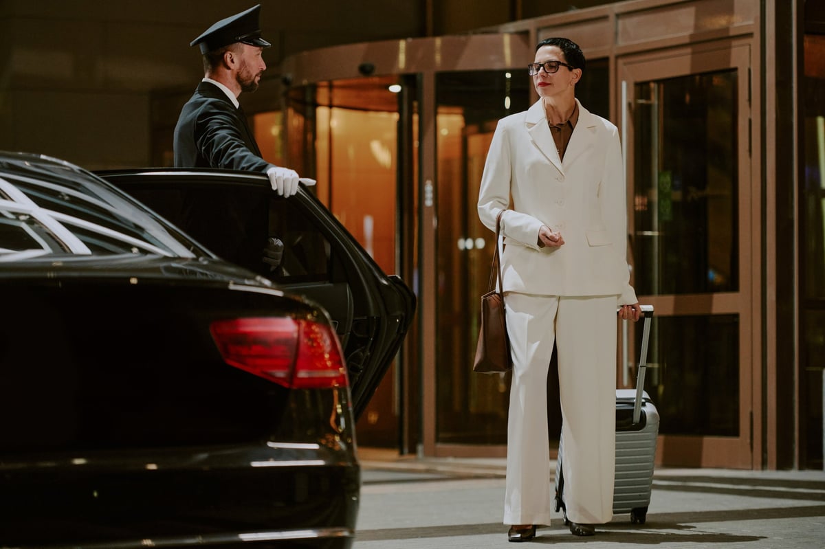 Woman standing outside hotel with suitcase and chauffeur opening car door