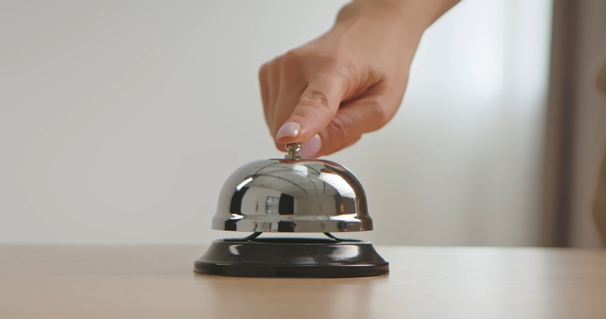 A close up of a hand pressing a shiny silver service bell placed on a wooden desk