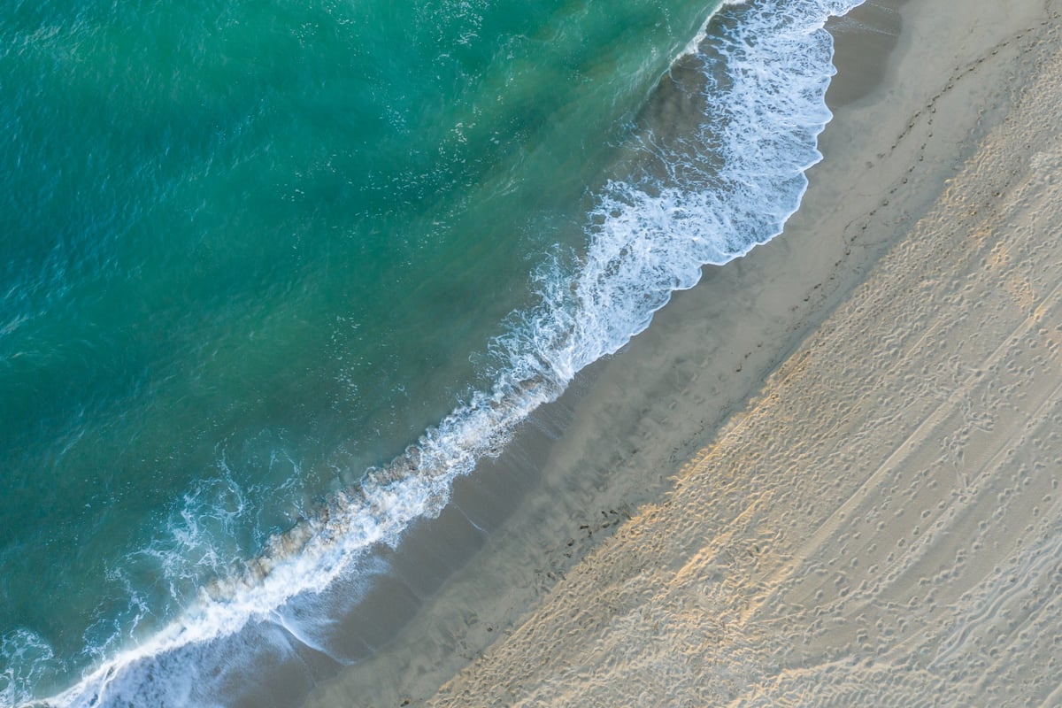 Aerial perspective of gentle turquoise waves rolling onto a smooth sandy beach, with visible footprints and soft textures under natural daylight