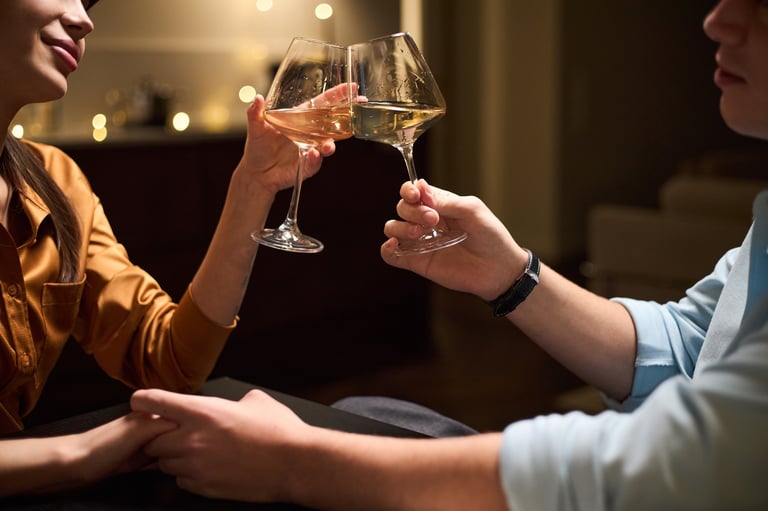 Couple clinking wine glasses while holding hands at table, smiling in romantic celebration