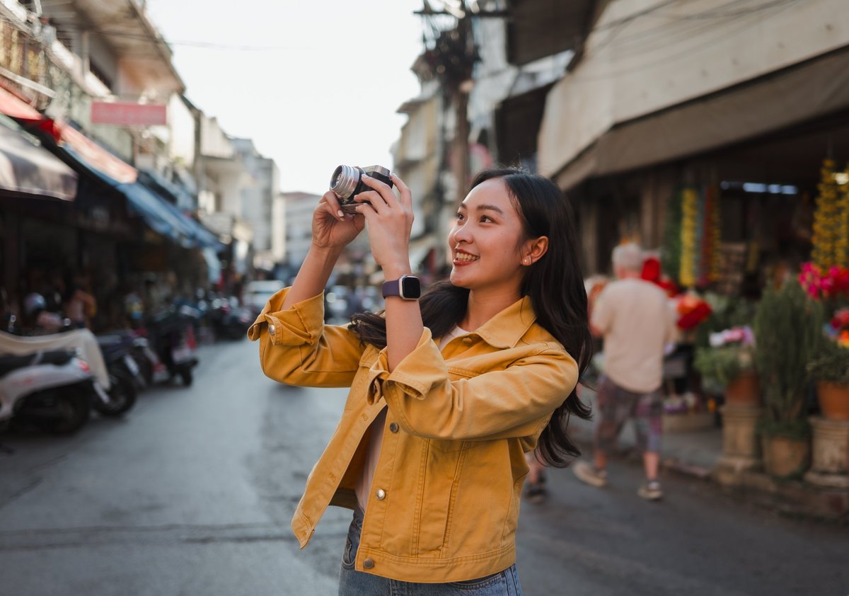 Young adult woman tourist wearing a yellow jacket and holding a retro camera, smiling while exploring an urban street