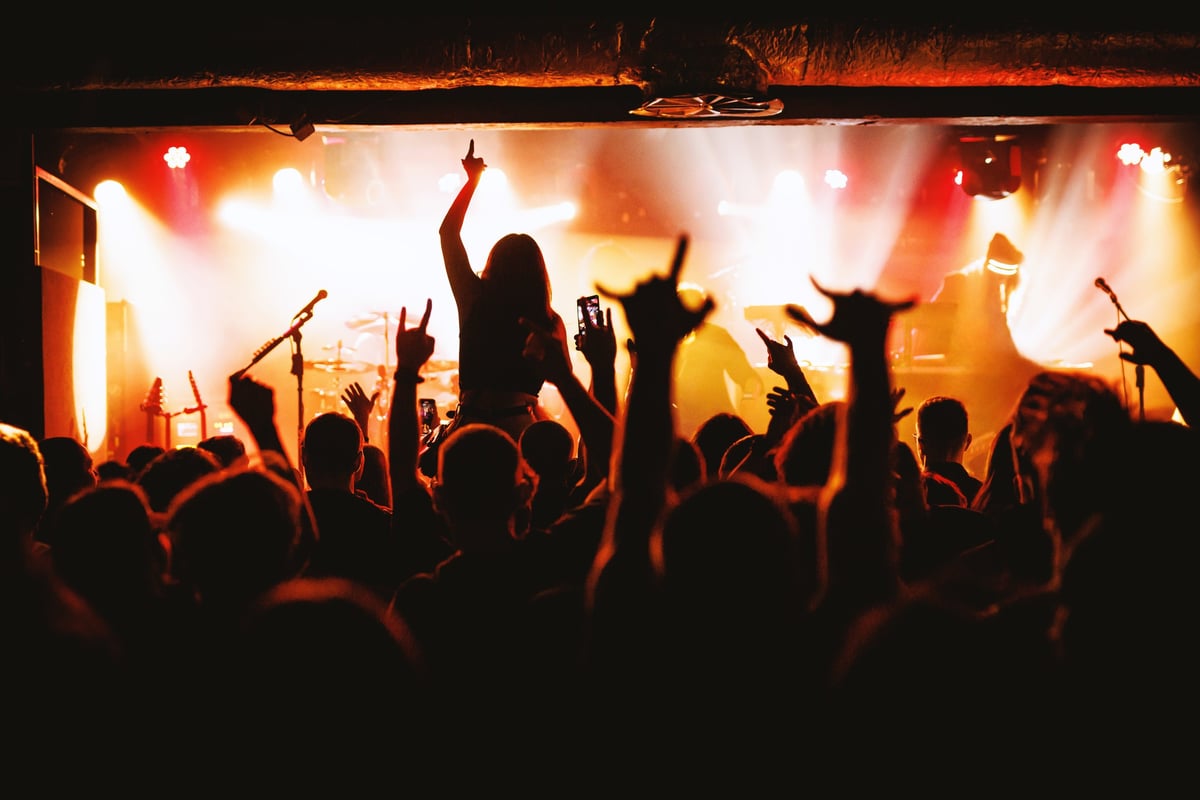 A girl sits on someone's shoulders in a concert crowd with her arm raised toward the bright stage during an intense live rock show