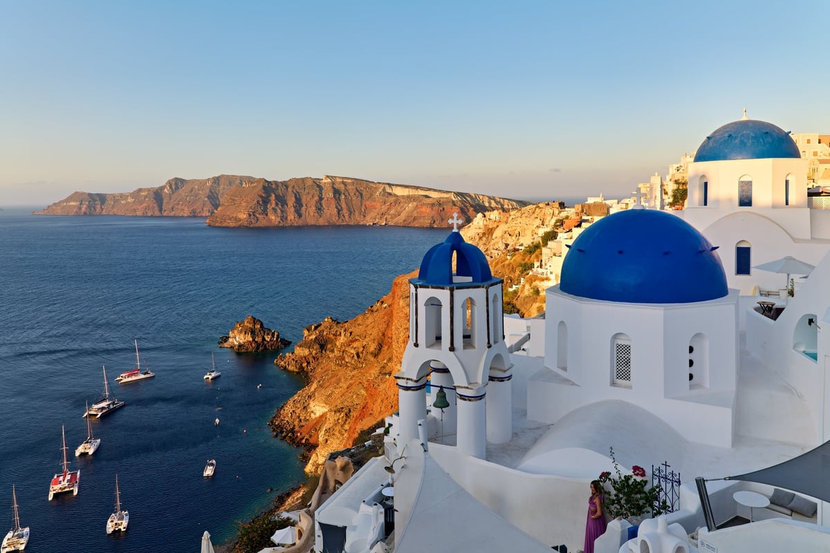 A woman enjoys the sunset view over the caldera in Oia, Santorini, Greece, with iconic blue-domed churches and boats in the Aegean Sea