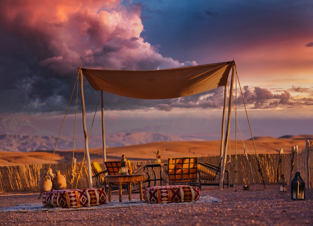 Outdoor seating area at a desert camp in Morocco with chairs, cushions, lanterns, and a canopy overlooking arid terrain at sunset