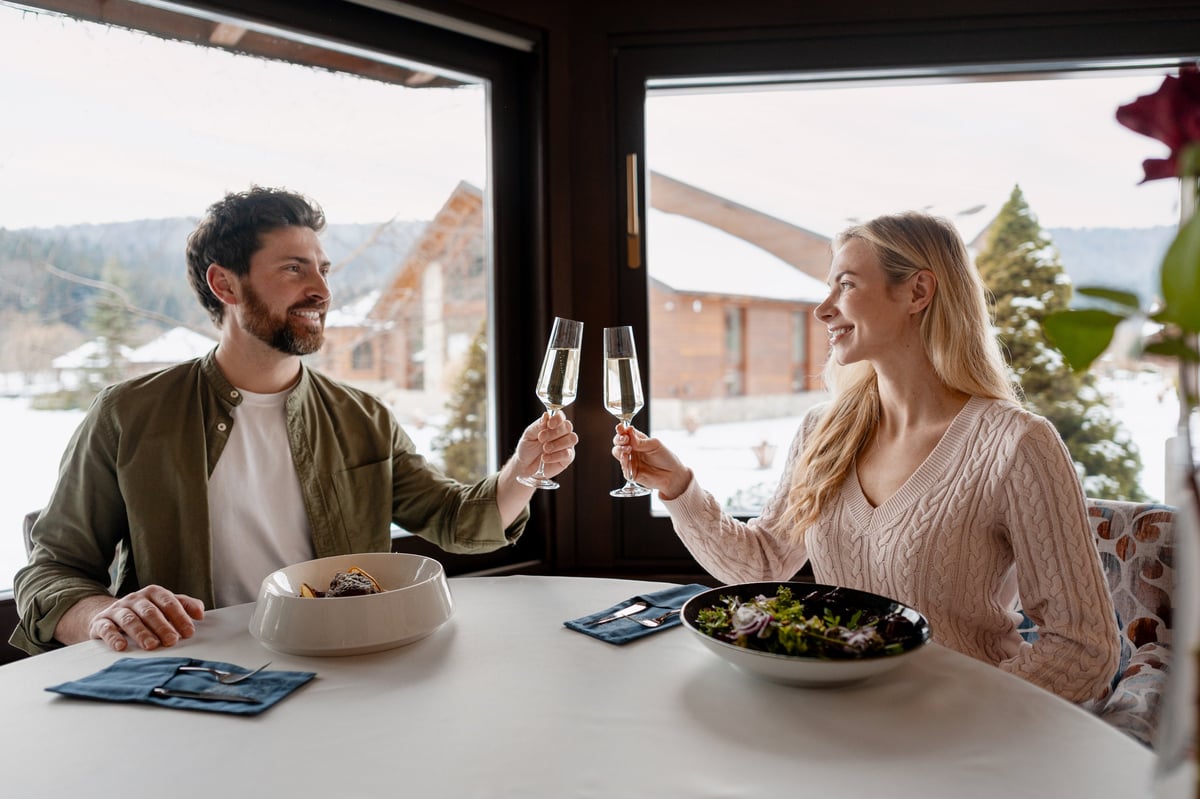 Happy couple toasting champagne glasses, enjoying meal at restaurant with snowy mountain view in the Alps