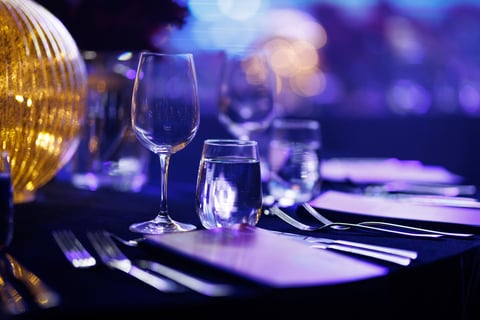 A crystal wine glass and a water tumbler sit on a dark tablecloth under blue and purple ambient lighting