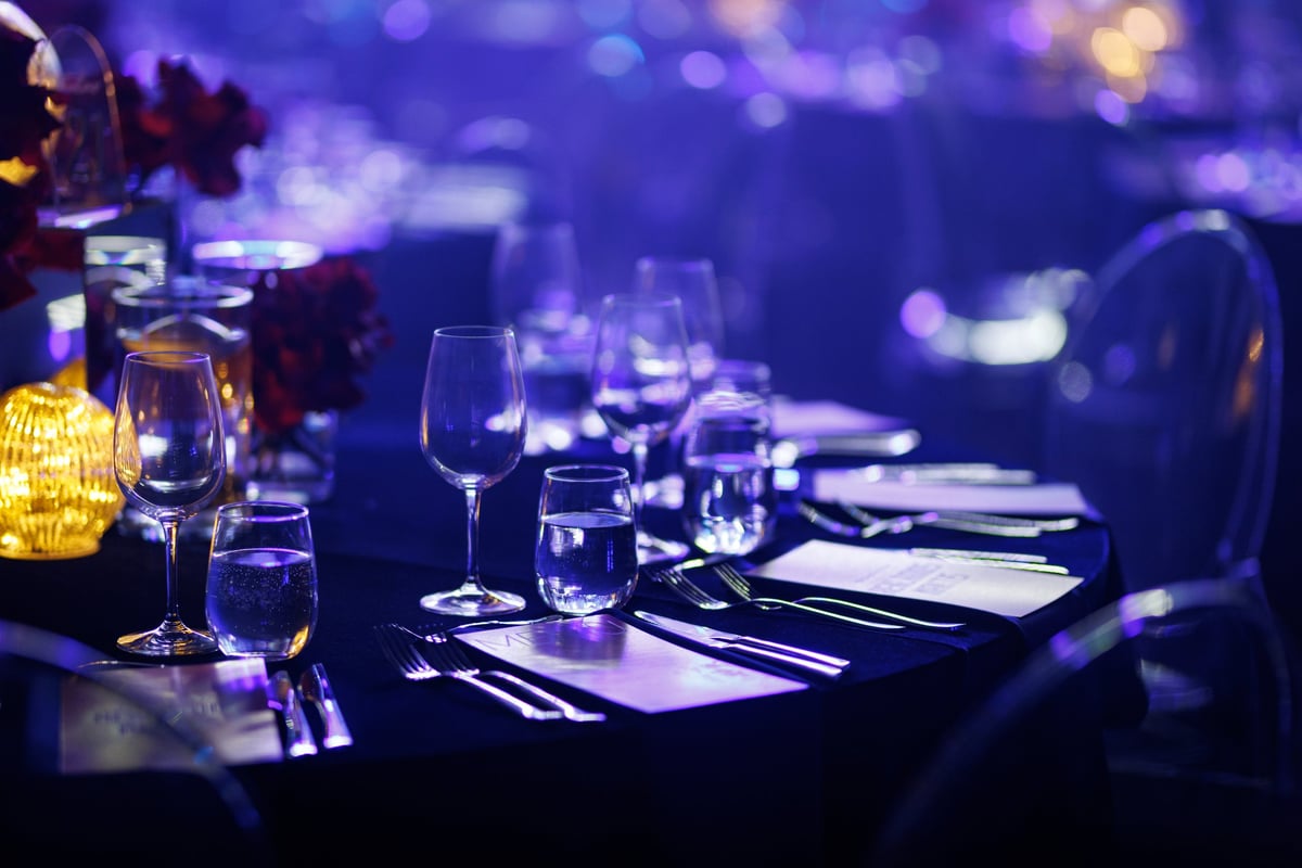 Formal dinner table with crystal glassware, polished cutlery, and menus under atmospheric blue stage lights