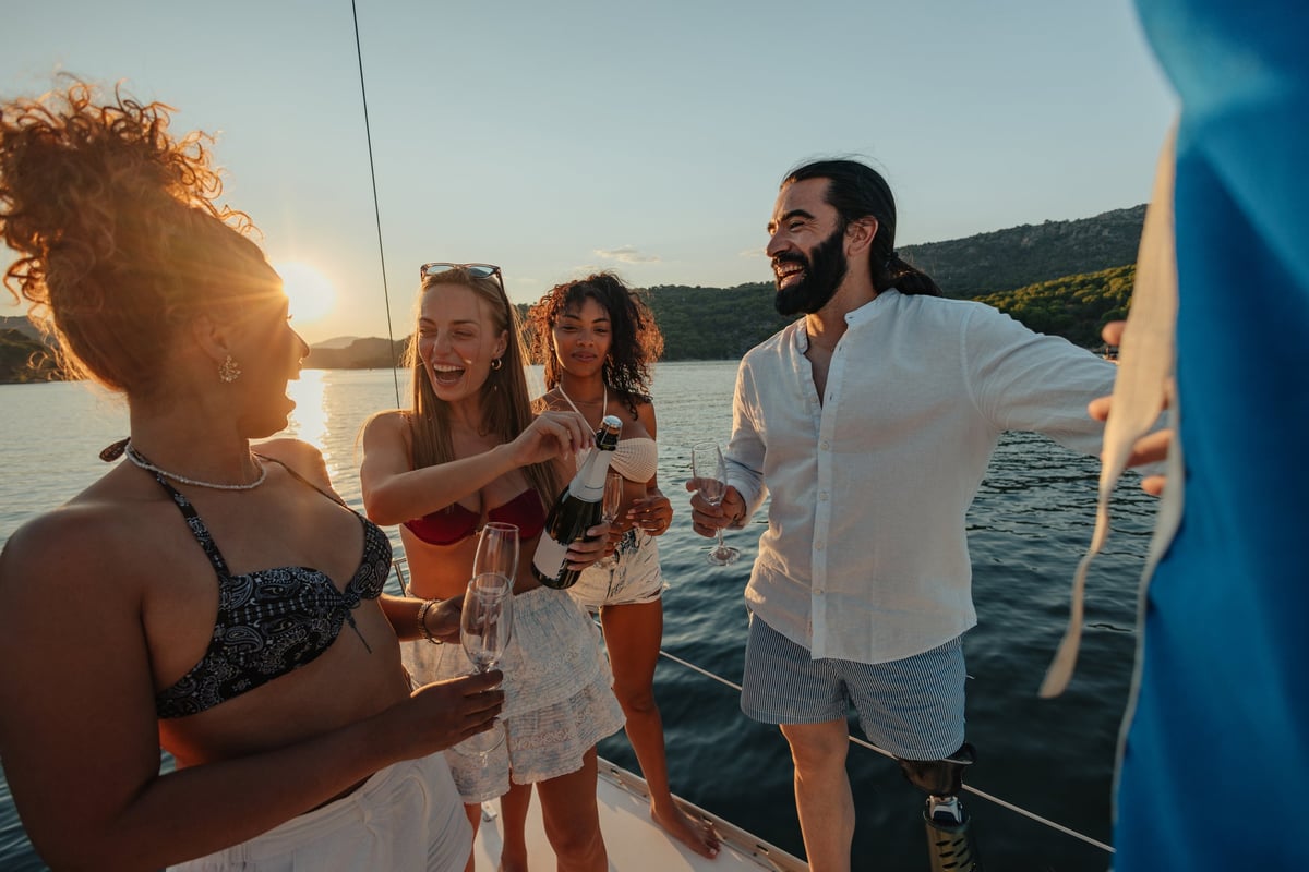 A diverse group of four friends laugh and open champagne aboard a sailing boat at sunset