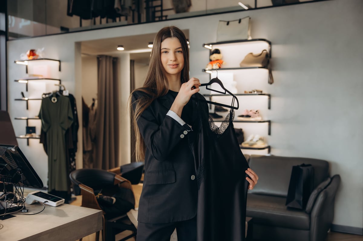 Young woman smiling, holding a black lace dress on a hanger in a fashion boutique, enjoying the shopping experience