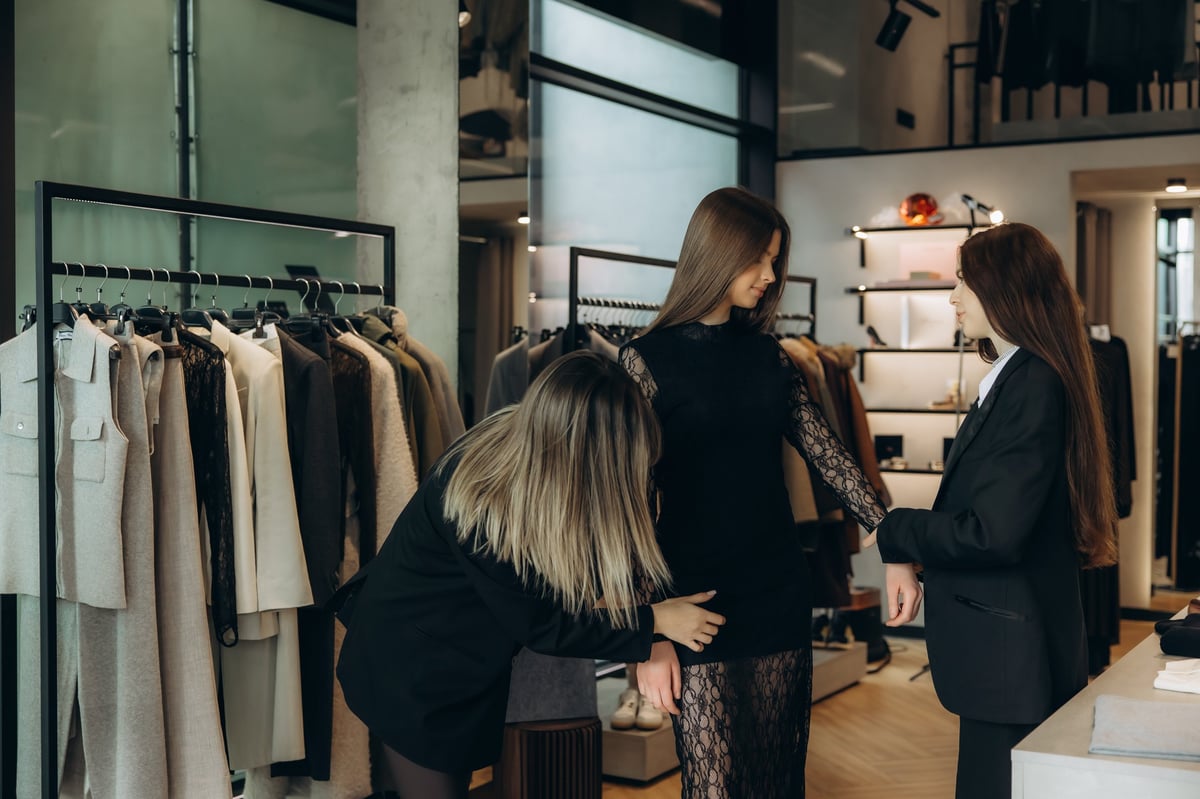 Stylist assisting a young woman trying on a black dress in a luxury fashion boutique with another assistant nearby