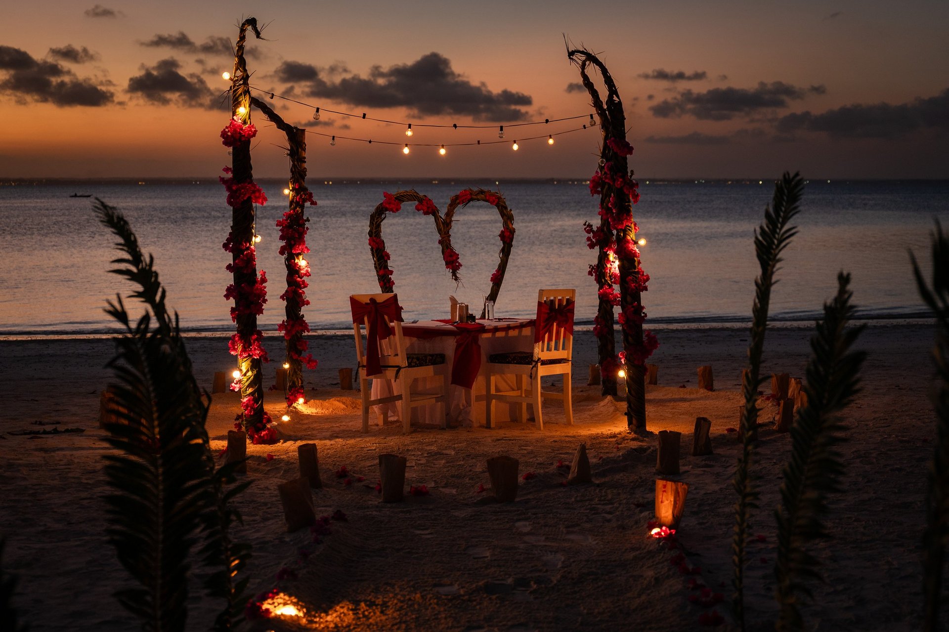 Romantic dinner table for two set on a beach at sunset with flower arches and decorative lights
