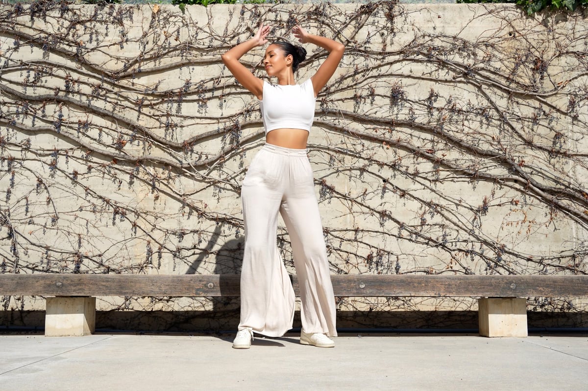 Woman posing against vine covered wall wearing white crop top and beige trousers, relaxed confident stance, sunlit textured concrete background, minimal bench