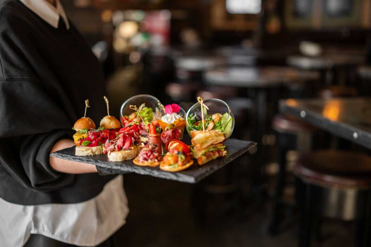 A waiter holds a slate tray of gourmet appetizers in a dimly lit, upscale bar or restaurant