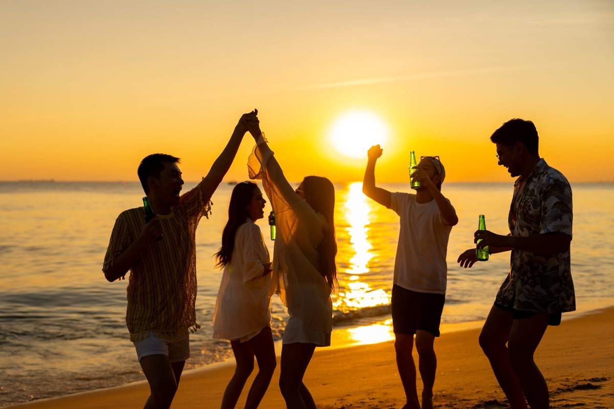 Group of diverse friends dancing and drinking on the beach at sunset celebrating life