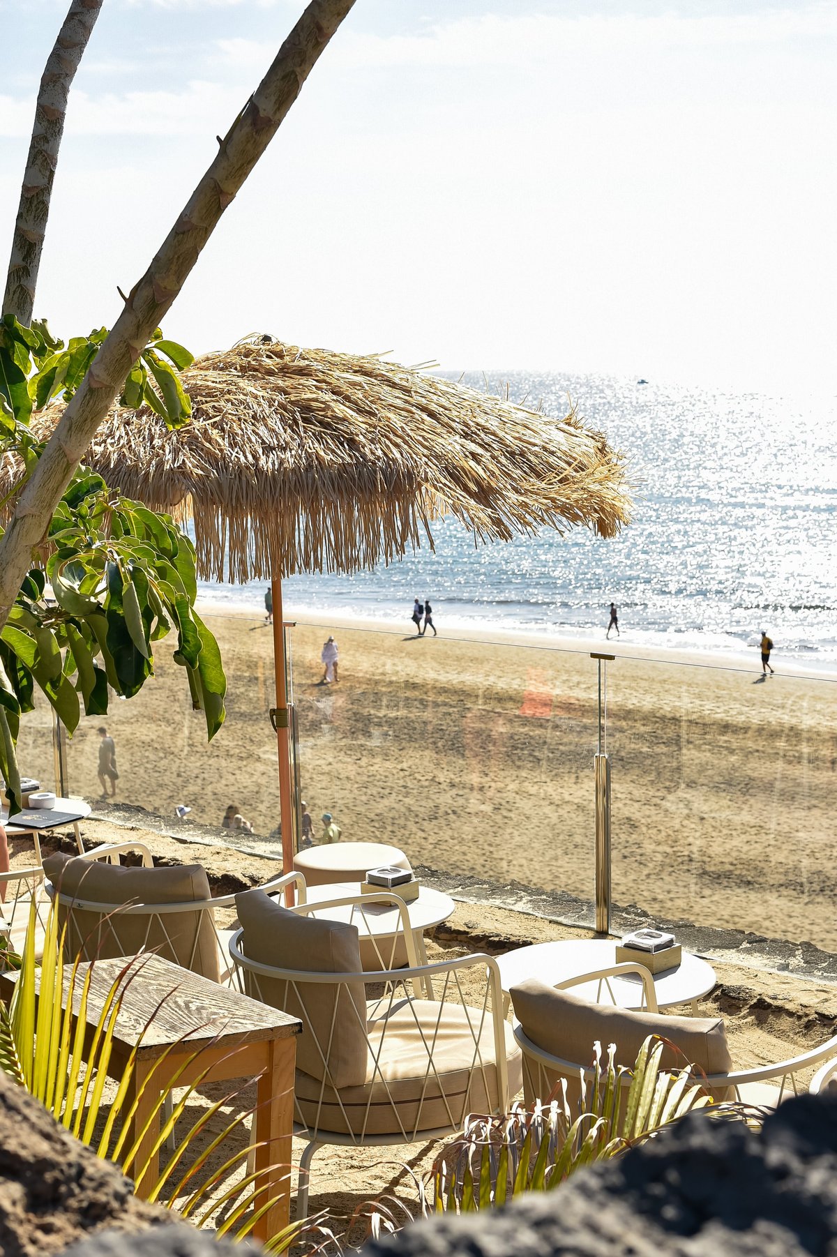 A straw sun umbrella shades a cafe terrace with modern lounge chairs by the ocean