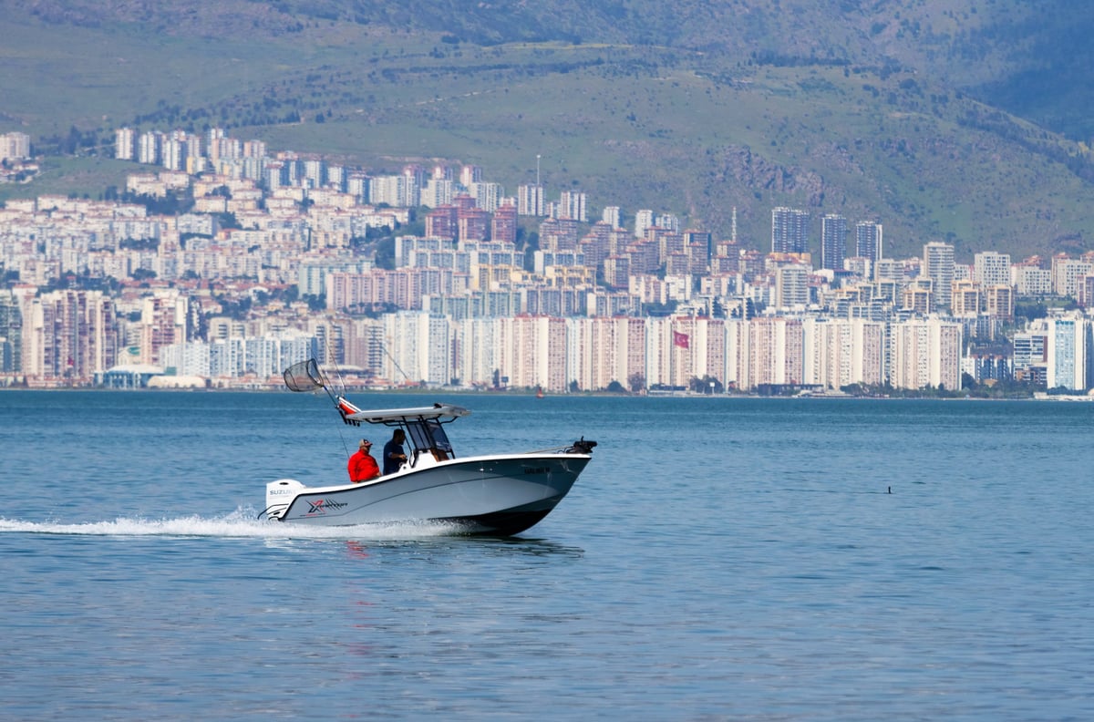 A grey speed boat with two people cruising on the calm blue waters of the bay with the city skyline and mountains in the background.