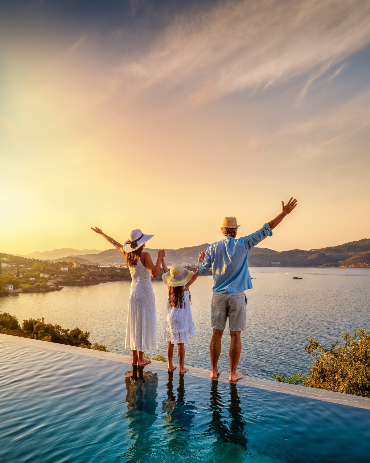 A happy family on summer holidays stands by the swimming pool and enjoys the beautiful sunset behind the ocean