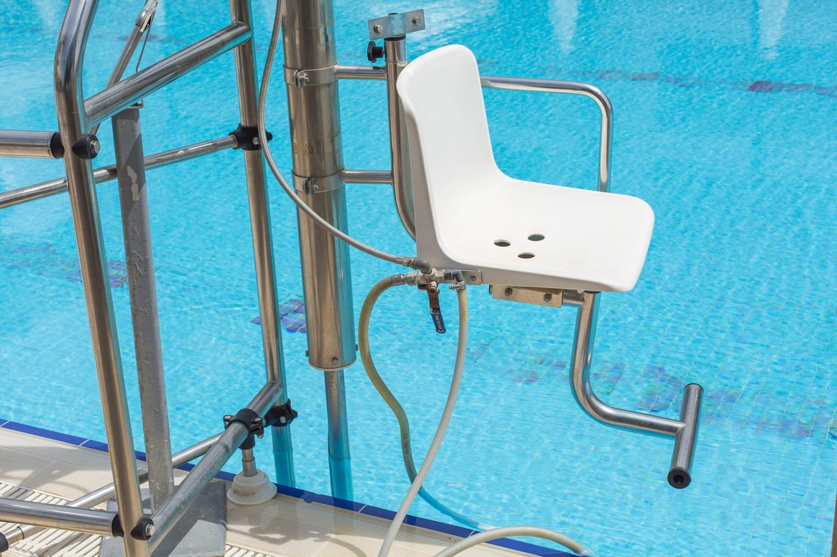 Poolside accessibility lift with white seat and metal hydraulic mechanism installed at edge of blue swimming pool, equipment designed for disabled access and safe water entry, inclusive environment concept