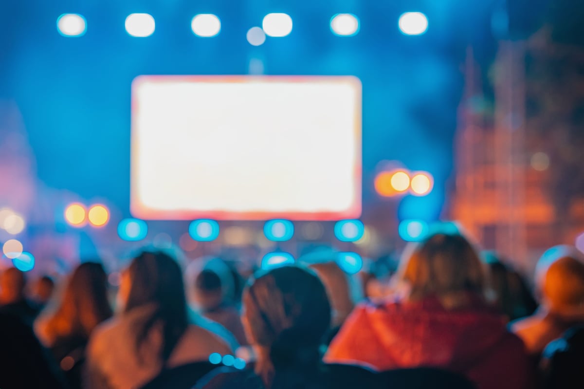 Rear view of blurred crowd watching large screen at night outdoor concert with lights