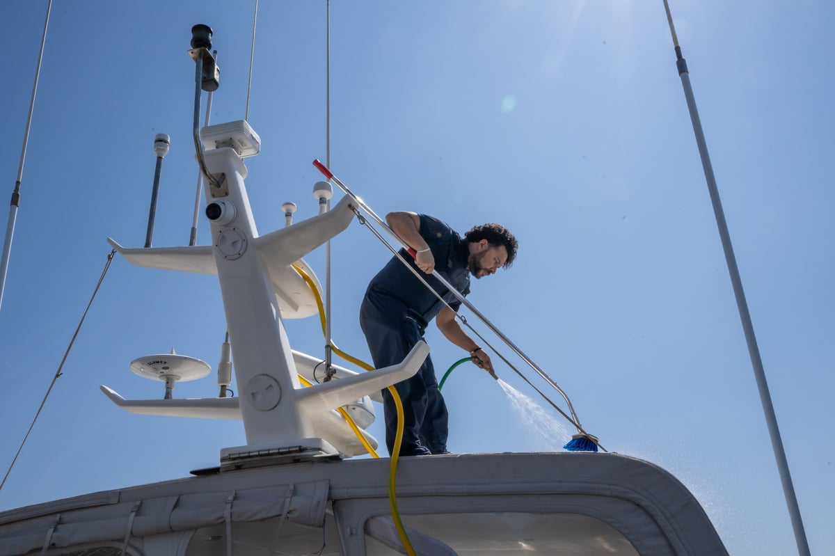 Professional yacht maintenance worker cleaning the upper deck of a luxury motor yacht under bright summer sunlight at a modern marina