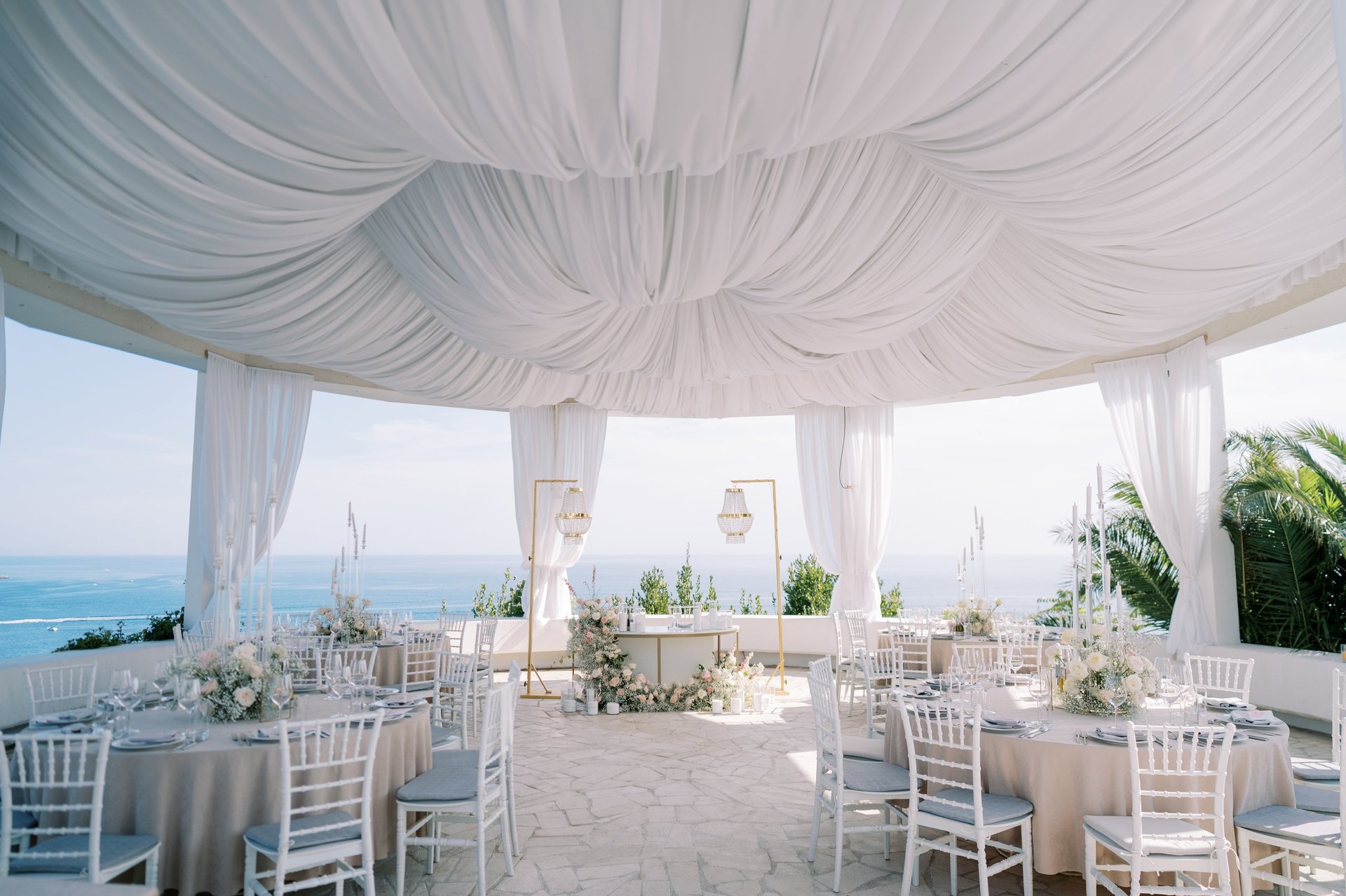 Beautifully decorated outdoor wedding reception under white canopy with ocean view, round tables, floral centerpieces